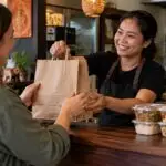 Friendly Thai takeaway counter conversation between customer and staff with unbranded paper bag and sealed meal containers