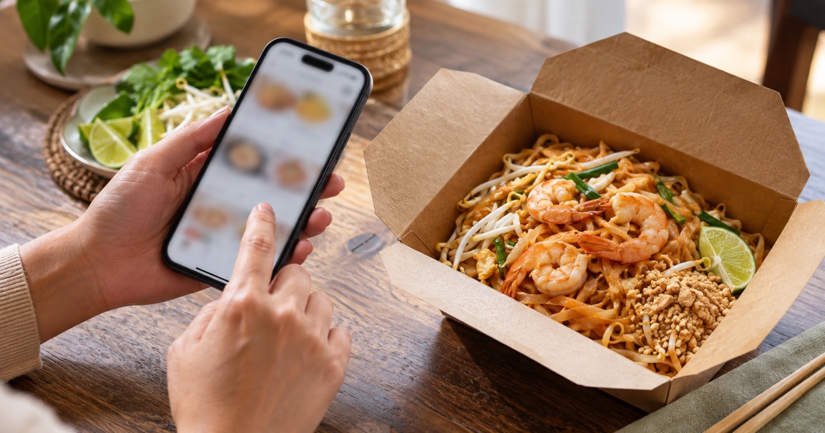 Hands holding smartphone beside Thai Pad Thai takeaway meal, screen fully blurred, simple online order concept