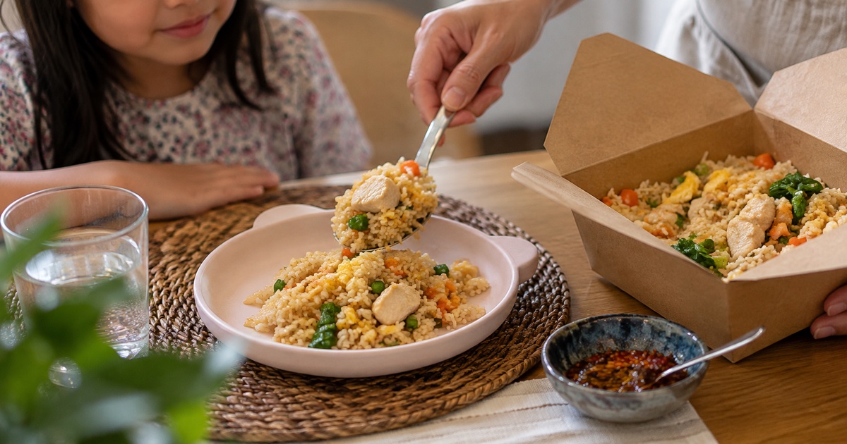 Close-up serving spoon transferring Thai fried rice from takeaway box to a small child plate, chilli condiment separate